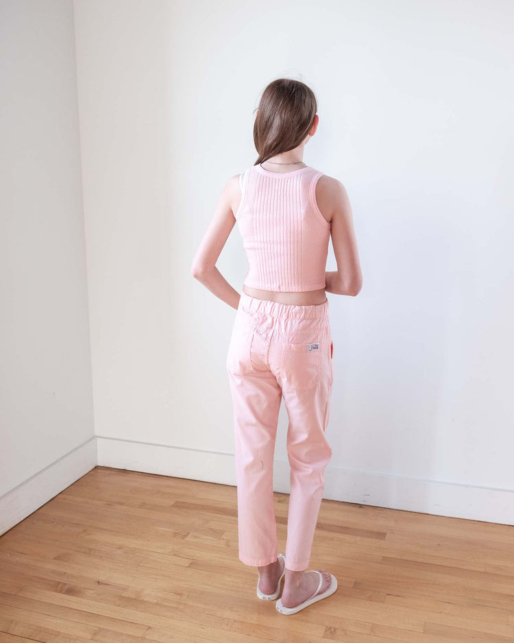 A person with long brown hair, barefoot, faces a white wall wearing a light pink sleeveless crop top and sunchild’s mustang pants in baby on a wooden floor.