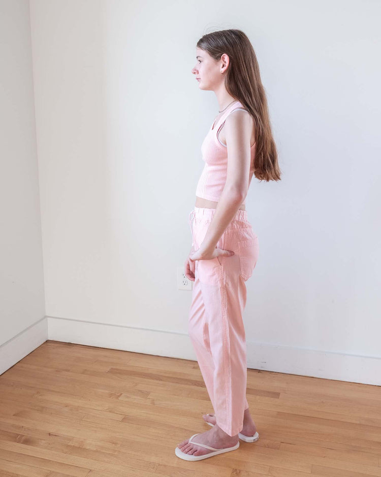 A young woman with long brown hair stands sideways in a light room, wearing a light pink tank top, Sunchild’s mustang pants in baby (100% cotton, straight leg), and white flip-flops.