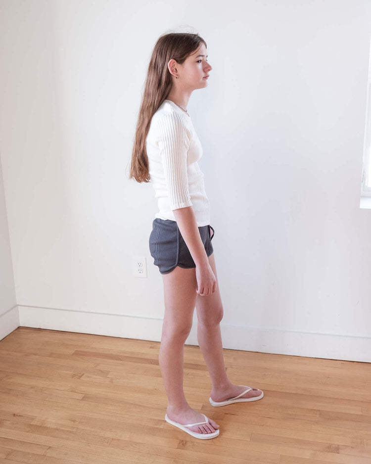 A young person with long brown hair, wearing the sunchild puebla tee in off white, dark shorts, and white flip-flops stands sideways in a simple room with light-colored walls and wooden floors.