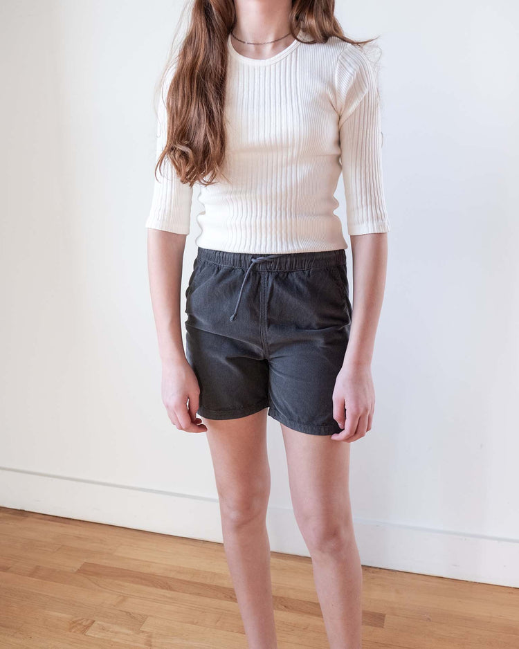 A person with long brown hair wears a white ribbed top and Sunchild's Hunter shorts in caviar, featuring a drawstring waistband. They stand indoors on a wooden floor against a white wall.