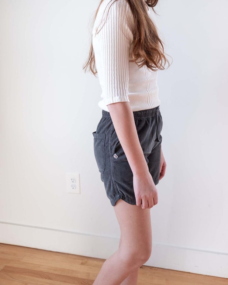 A person with long brown hair stands indoors, wearing a white ribbed three-quarter sleeve top and sunchild hunter shorts in caviar. The setting includes a white wall, power outlet, and wooden floor.