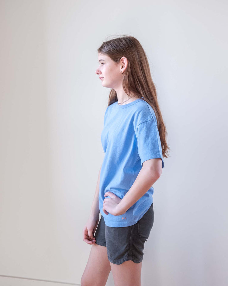 A young Sunchild with long brown hair stands sideways, wearing a light blue T-shirt and Sunchild hunter shorts in caviar with a drawstring waistband, looking ahead with one hand on their hip against a plain light-colored wall.