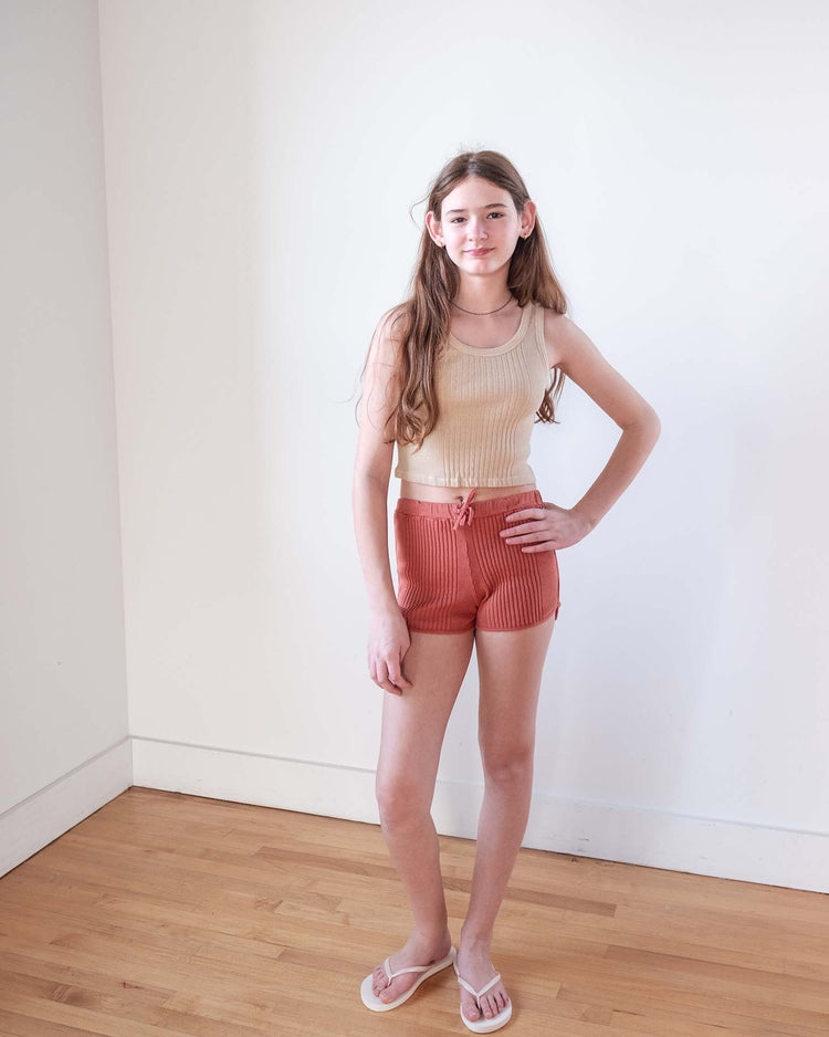 A young person with long brown hair stands indoors against a white wall, wearing the Sunchild cruz tank in craie, high-waisted salmon-colored shorts, and flip-flops on a wooden floor.