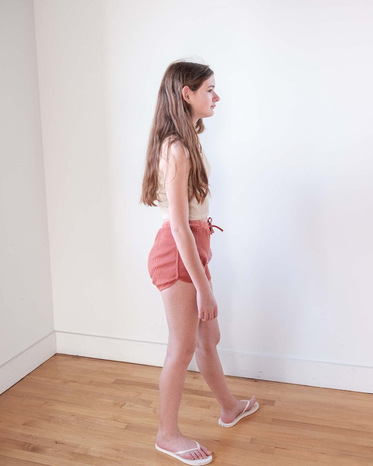 A teenage girl with long brown hair wears a sleeveless top and sunchild rio shorts in cacao with a drawstring waist, standing in profile against a white wall and wooden floor, looking forward in white flip-flops.