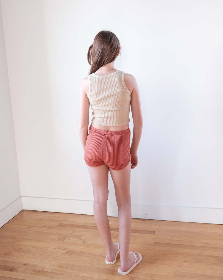 A girl with long brown hair stands indoors, facing away from the camera. She wears a beige sleeveless top, white sandals, and sunchild's rio shorts in cacao, featuring a 100% cotton drawstring waist, on a wooden floor by a white wall.