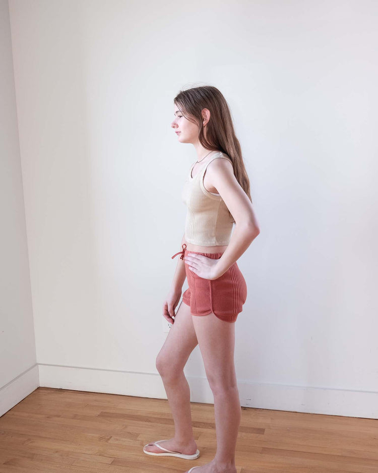 A young woman with long hair stands in profile against a white wall, wearing a beige tank top and sunchild rio shorts in cacao (100% cotton, drawstring waist), paired with white sandals on a wooden floor.