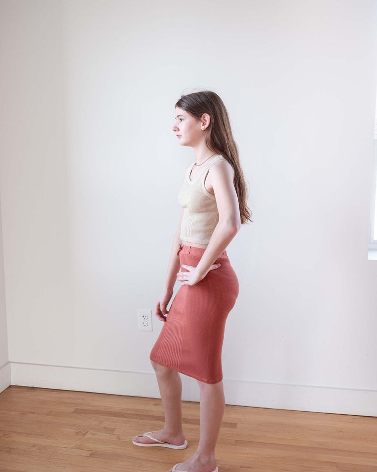 A young woman with long brown hair stands in profile indoors, wearing a sunchild cruz tank in craie, a high-waisted rust-colored skirt, and sandals. She faces left against a plain white wall with wooden flooring.