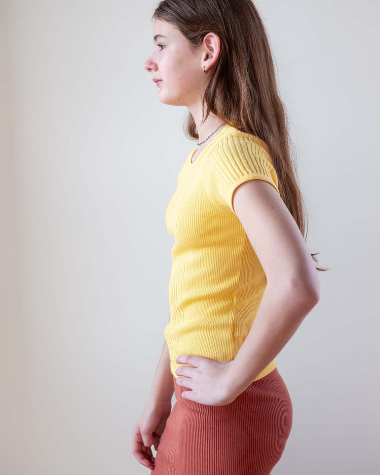 A young woman with long brown hair stands in profile, wearing the sunchild loutro tee in napoli—a fitted short-sleeve cotton top—and rust pants, set against a plain light background.