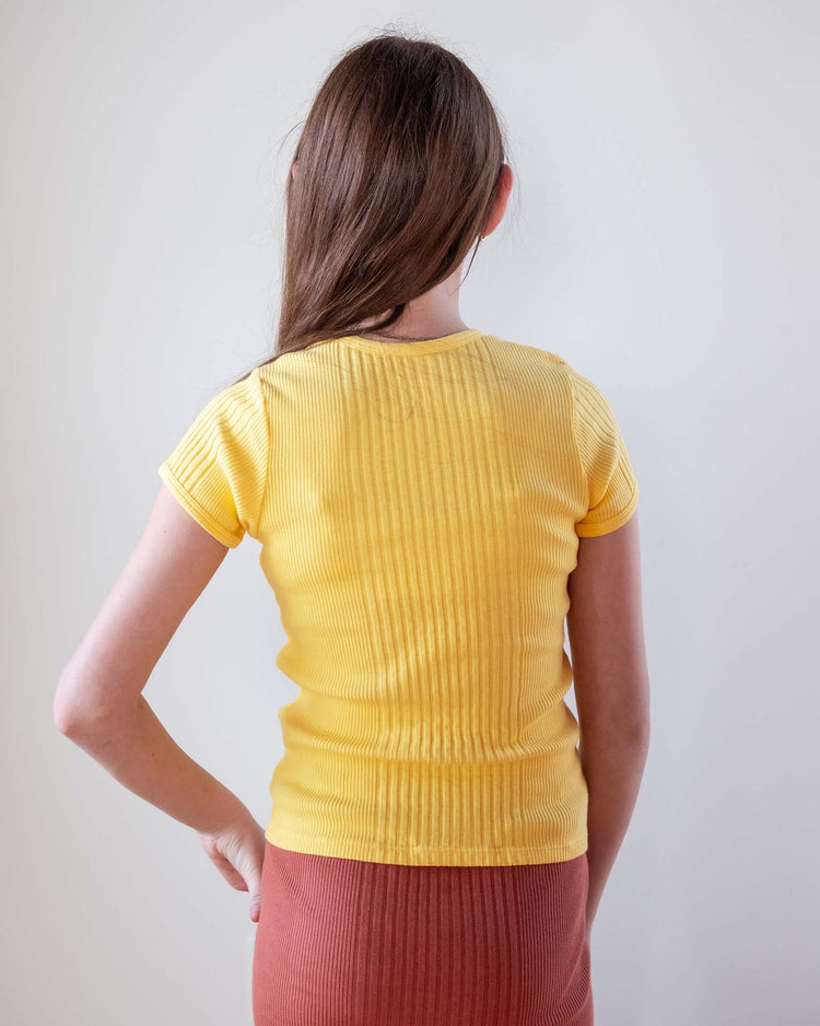 A woman with long brown hair stands facing away, wearing the sunchild loutro tee in napoli yellow and a rust-colored skirt, her left hand on her hip against a plain light background.