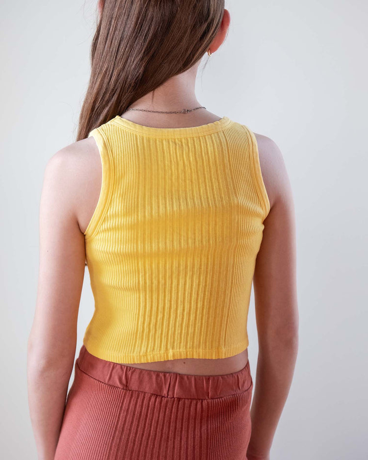 A person with long brown hair, seen from behind, wears a sunchild cruz tank in napoli and high-waisted rust-colored ribbed pants against a plain light background.