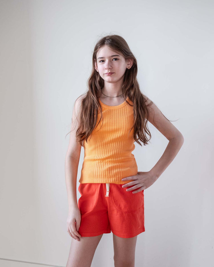 A girl with long brown hair stands against a white wall, wearing an orange sleeveless top and Sunchild merida shorts in tangerine with a drawstring waist, her hand on her hip as she looks forward with a neutral expression.