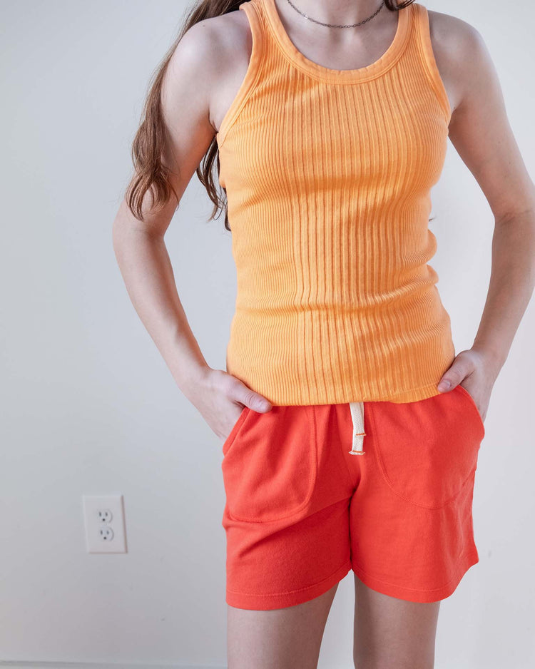 Wearing a ribbed orange tank top and sunchild merida shorts in tangerine with a drawstring waist, a person stands against a plain white wall, hands in their pockets. Their head is out of frame.