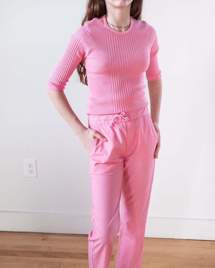 A person stands indoors by a white wall wearing the sunchild puebla tee in glamour—a light pink ribbed cotton style—paired with matching pants. One hand is on their hip; a power outlet and wooden floor are visible in the background.