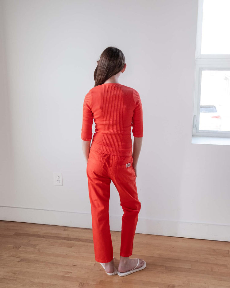 A person with long brown hair stands facing away, wearing the sunchild puebla tee in tangerine. They're by a white wall and window, on a wooden floor, hands at their sides.