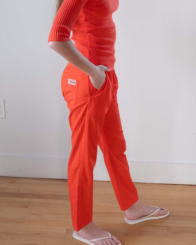 A person stands indoors with hands in pockets, wearing a bright orange ribbed top, Sunchild mustang pants in tangerine, and white flip-flops. Only their torso and legs are visible.