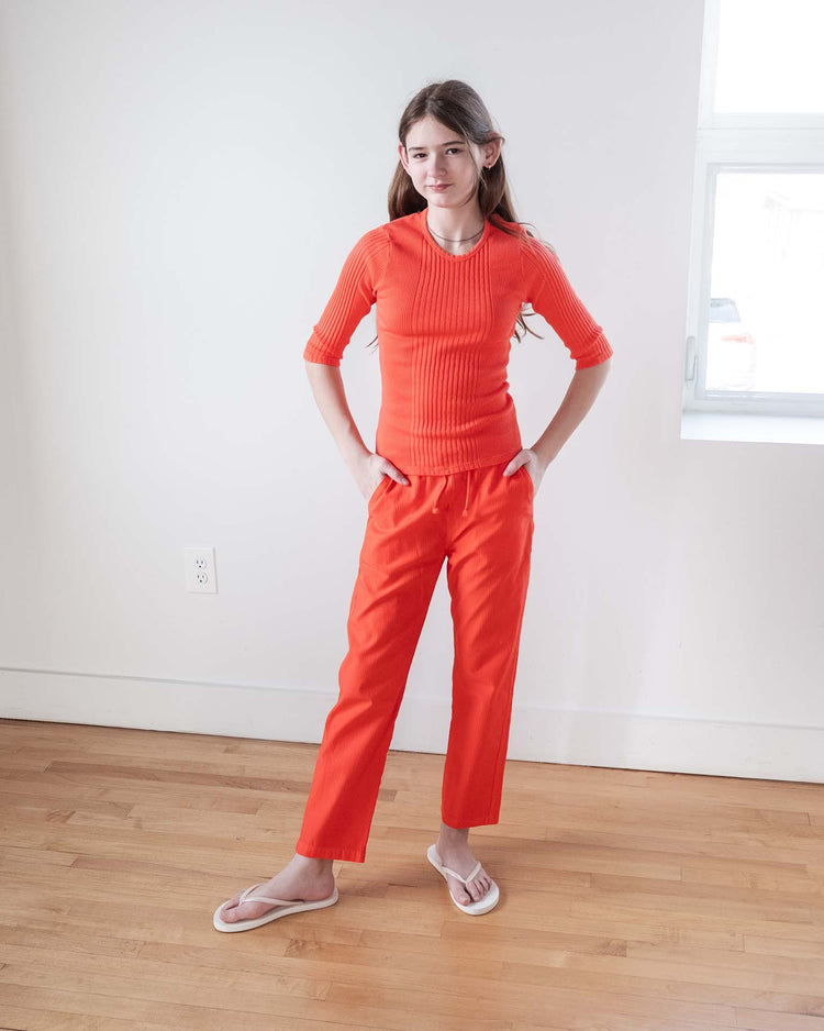 A young person stands indoors on a wood floor, hands in pockets and a slight smile, wearing the sunchild puebla tee in tangerine with matching orange pants and white flip-flops. A window appears in the background.