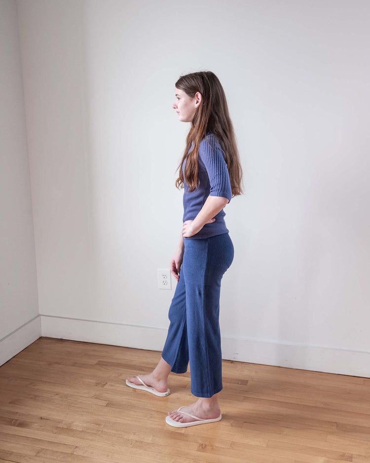 A woman with long brown hair stands in profile in a minimalist room, wearing the sunchild puebla tee in obscur, blue cropped pants, and white flip-flops, with her left hand resting on her hip.