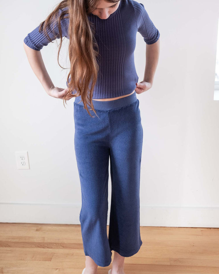 A person with long brown hair wears the Sunchild Puebla tee in obscur and loose blue pants, standing barefoot on a wooden floor near a white wall, embracing relaxed Marseille-inspired style.
