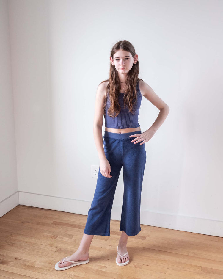 A young girl with long brown hair stands indoors against a white wall, wearing sunchild's bluff pants in obscur, a blue crop top, and white flip-flops. She poses with one hand on her hip and maintains a neutral expression.