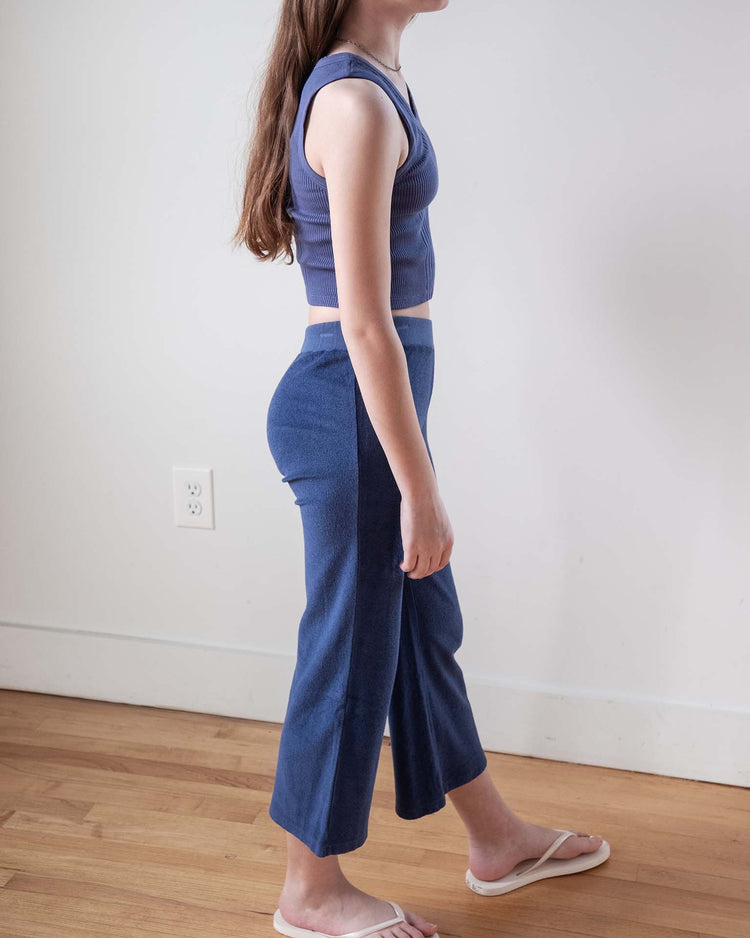 A person with long brown hair stands sideways against a plain white wall and hardwood floor, wearing a sleeveless blue crop top, sunchild bluff pants in obscur, and white flip-flops.
