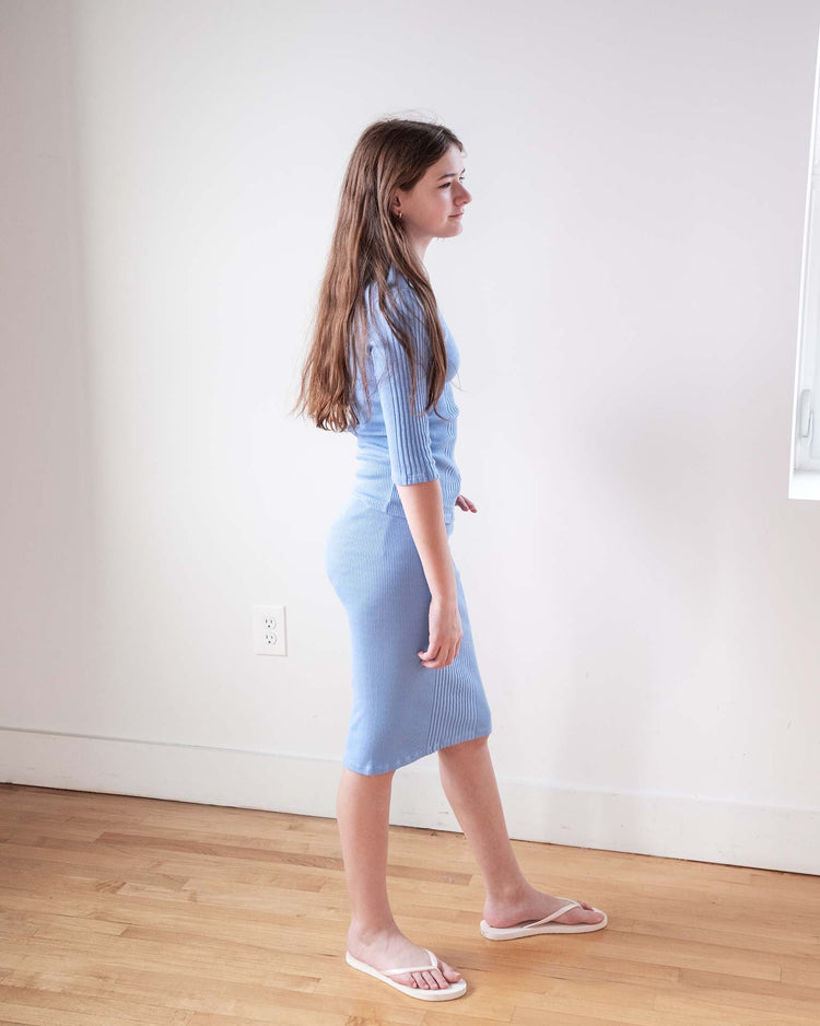 A young woman with long brown hair stands indoors on a wooden floor, wearing the sunchild siuna skirt in mare and white sandals, facing sideways in front of a plain white wall.