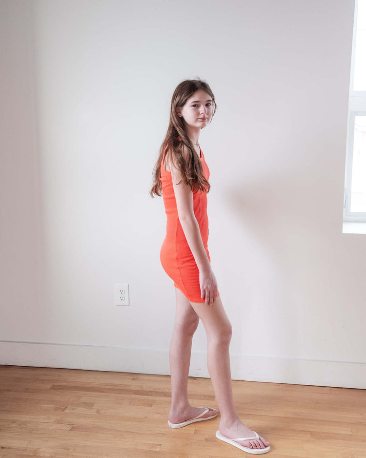 A young woman with long brown hair stands indoors against a white wall, wearing the sunchild pipa dress in tangerine and white sandals. She faces sideways, looking at the camera as daylight streams in from a window on the right.