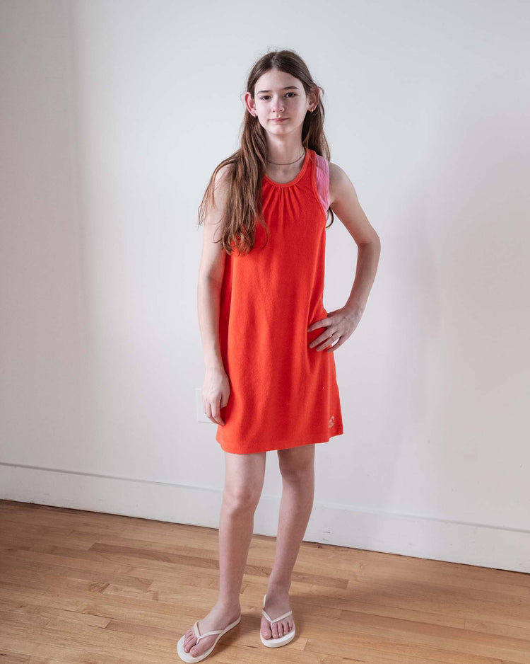 A young person with long brown hair poses indoors on a wooden floor in true Sunchild style, wearing the sunchild bilbao dress in tangerine—a sleeveless 100% cotton tank dress—with white flip-flops against a plain white wall.