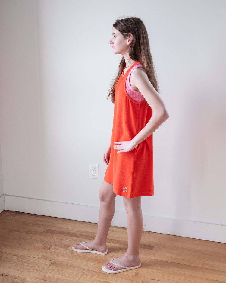 A young person with long hair stands sideways in a room, wearing the sunchild bilbao dress in tangerine over a pink top and flip-flops. The plain white wall and wooden floor create a simple background.