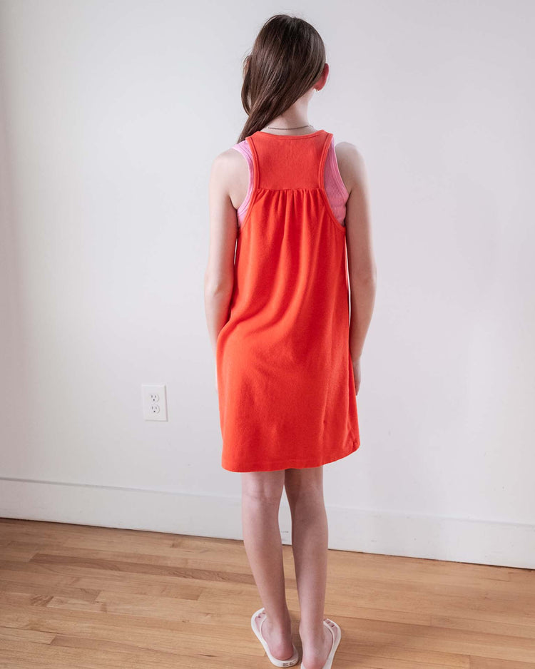 A young girl with light skin and brown hair stands indoors on a wooden floor, facing a white wall, wearing the Sunchild bilbao dress in tangerine over a light pink top and paired with white flip-flops.