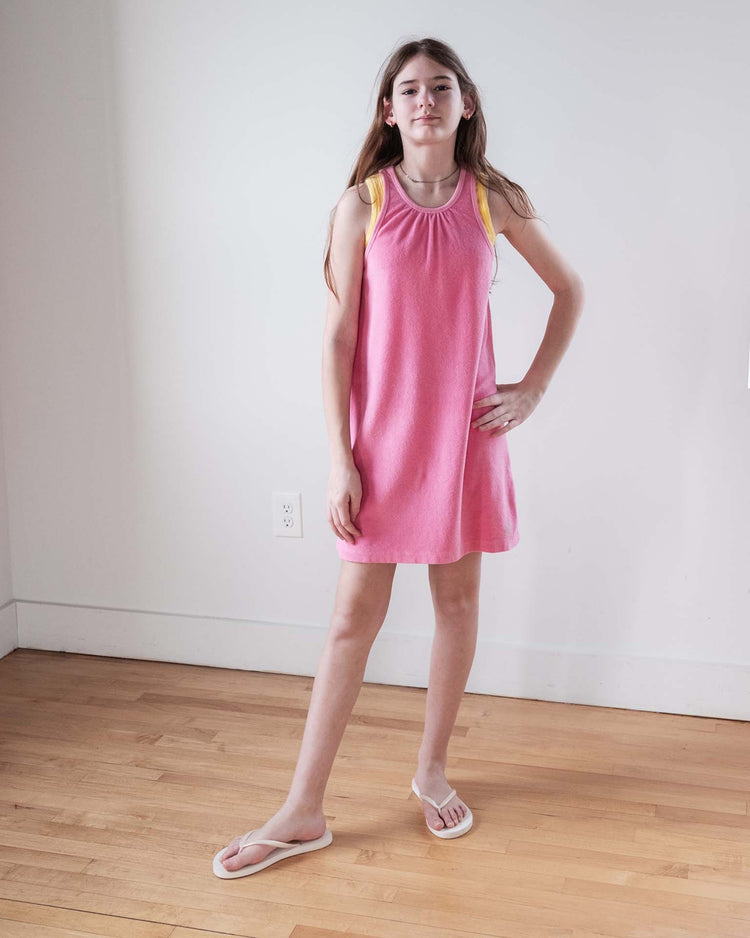 A young girl with long hair stands indoors on a wooden floor, wearing the sunchild bilbao dress in glamour and white flip-flops. She poses with one hand on her hip and a neutral expression.