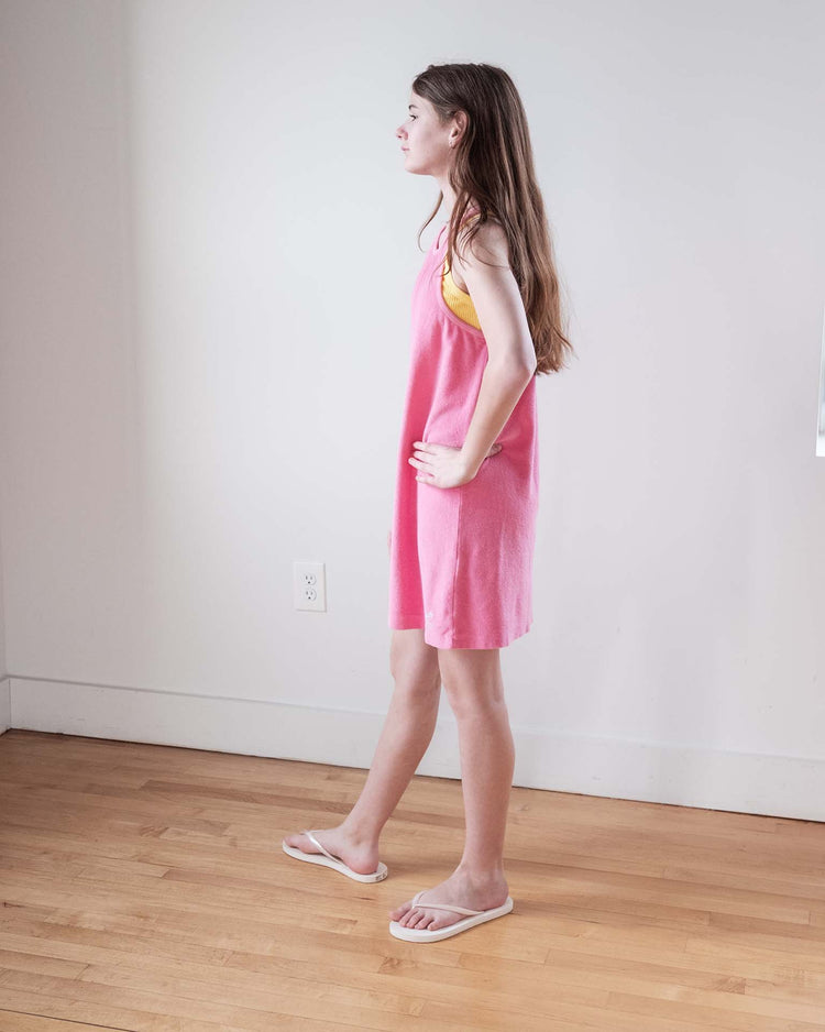 A young woman with long brown hair stands sideways in a room with white walls and a wooden floor, wearing the sunchild bilbao dress in glamour. She has her hands on her hips and looks ahead.