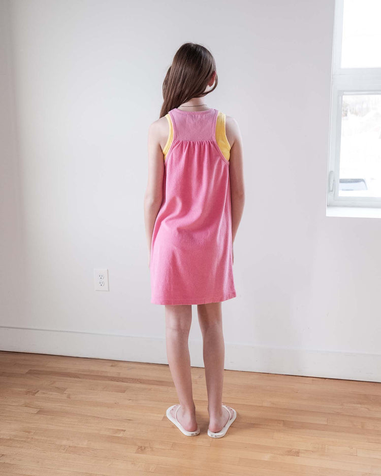 A young girl with long brown hair stands indoors facing away, wearing the sunchild bilbao dress in glamour and white flip-flops. She is on a wooden floor near a white wall and a window with daylight streaming in.