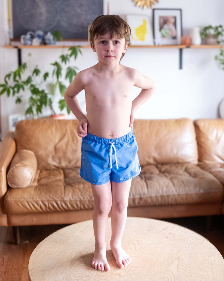 A young boy in sunchild bahia board shorts in mare stands barefoot on a round wooden table in a living room, with a tan leather sofa, plants, and shelves with framed photos in the background.