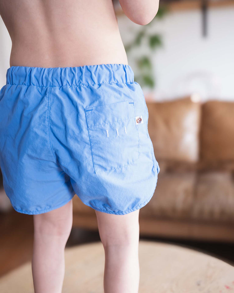 A child in light blue sunchild bahia board shorts in mare stands on a round wooden table indoors. The upper body is out of frame, with a brown sofa and plants softly blurred in the background.