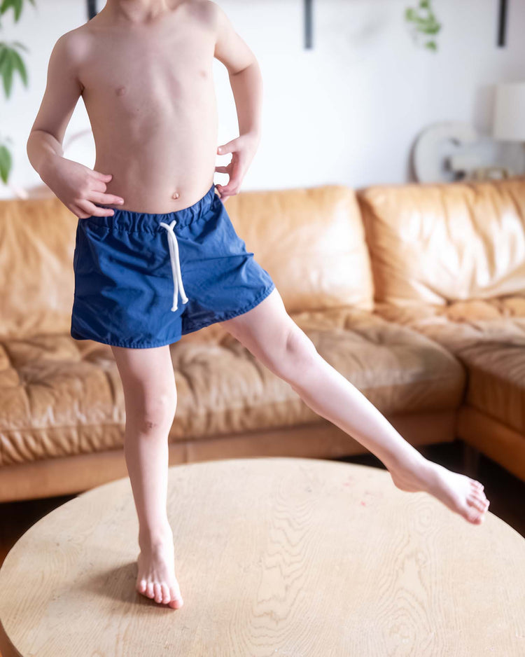 A shirtless child wearing sunchild's bahia board shorts in obscur stands on a round wooden table with one leg raised, a brown leather sofa visible in the living room background.