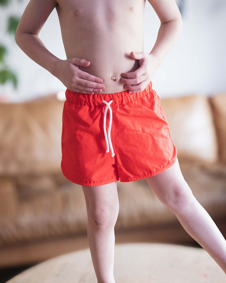 A child wears sunchild bahia board shorts in tangerine, standing indoors with hands on their bare stomach, one leg raised, and a couch in the background.