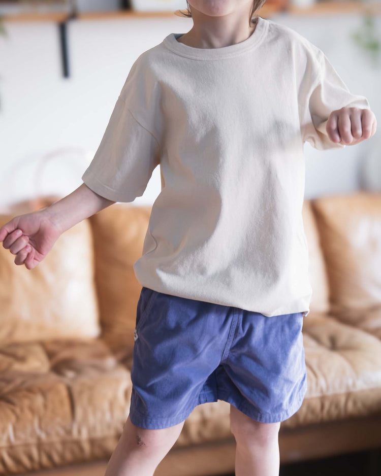 A child wears the sunchild kazan tee in craie and blue shorts, standing indoors before a tan leather couch with arms raised playfully. The child's face is partially out of frame.