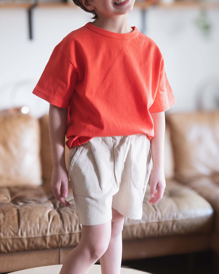 A Sunchild beams in a bright orange tee and sunchild merida shorts in craie, featuring a drawstring waist, while standing on a beige sofa indoors with softly blurred shelves and cozy household items in the background.