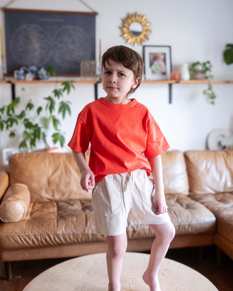 A young Sunchild in a bright orange shirt and sunchild's merida shorts in craie with a drawstring waist stands confidently on a round wooden table, with a brown leather sofa and plants in the background.