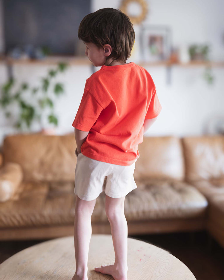 A young child with short brown hair stands barefoot on a round table, facing away in a cozy living room with a tan sofa and plants, wearing the sunchild kazan tee in tangerine and light shorts.