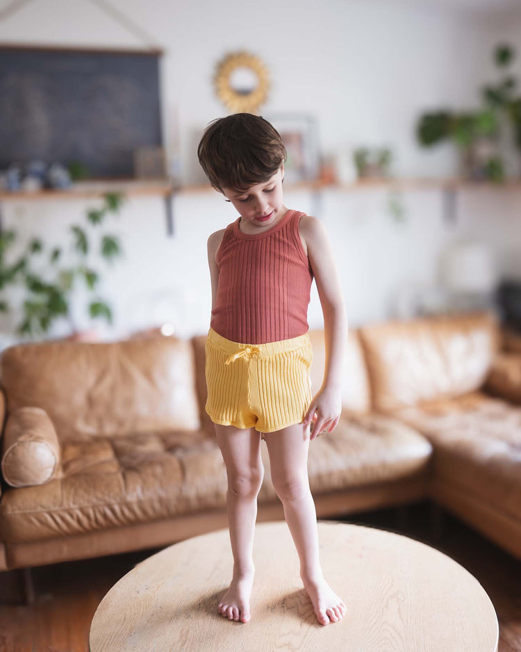A child in a sleeveless rust top and Sunchild's Rio shorts in Napoli with a drawstring waist stands barefoot on a round wooden table, with tan couches, shelves, and green plants in the living room background.