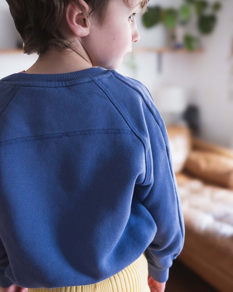A child faces away, wearing the sunchild douglas sweatshirt in obscur and yellow pants, standing in a cozy living room with a brown couch and green plants behind.
