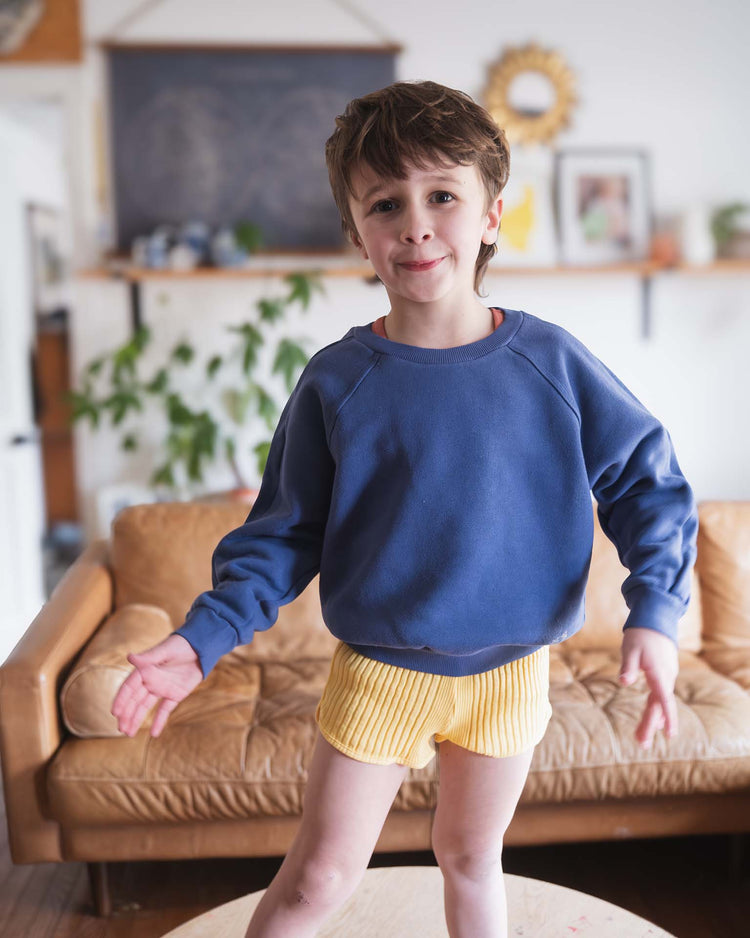 A young sunchild with short brown hair, wearing the sunchild douglas sweatshirt in obscur and yellow shorts, stands on a round table in a living room with a tan couch and plants in the background.