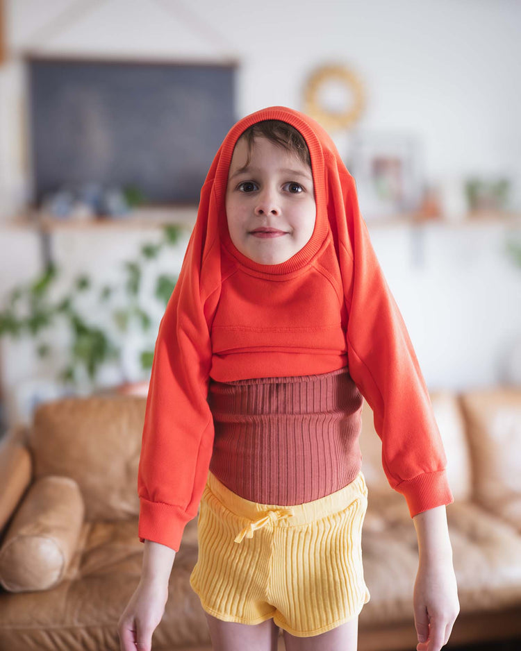 A young child stands indoors with the sleeves of the sunchild douglas sweatshirt in tangerine draped playfully over their head and torso, paired with yellow shorts. A couch and plants are visible in the background.