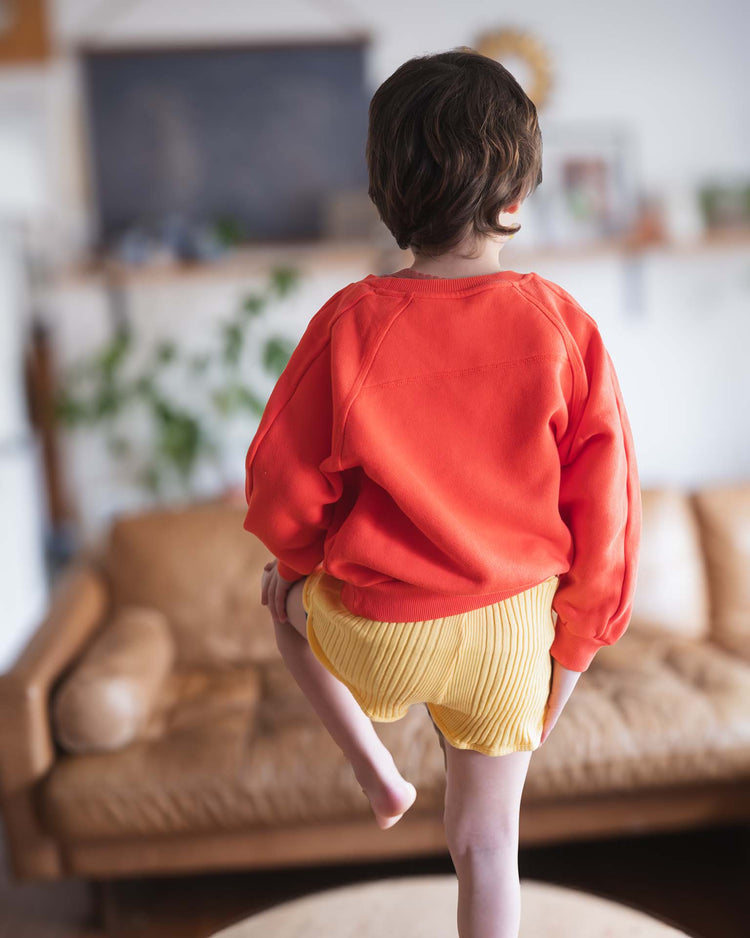 A Sunchild model with short brown hair wears the sunchild douglas sweatshirt in tangerine and yellow shorts, standing on a table indoors with one leg lifted. A brown couch and a blurred background are also visible.