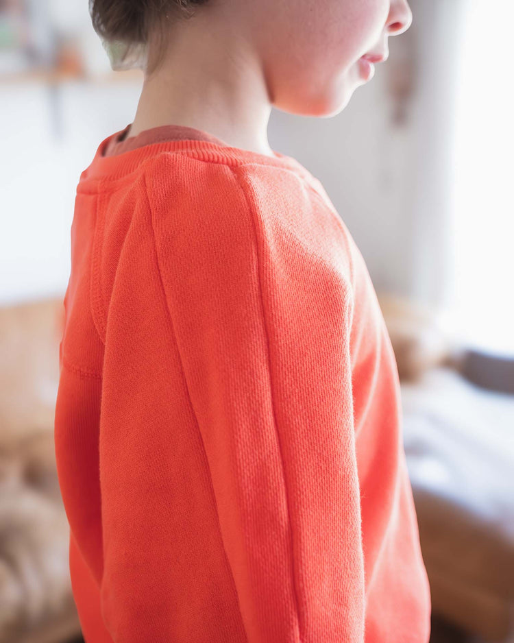 A close-up side view of a child wearing the Sunchild douglas sweatshirt in tangerine, shown with soft lighting and a blurred indoor background.