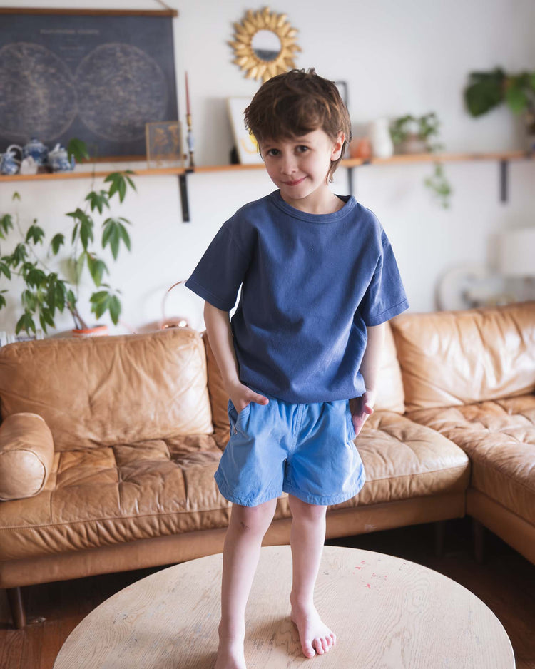 A young Sunchild wears sunchild's hunter shorts in mare—a light blue cotton style with a drawstring—paired with a loose blue t-shirt, standing barefoot on a round wooden table in a cozy living room with a tan leather sofa and plants behind him.