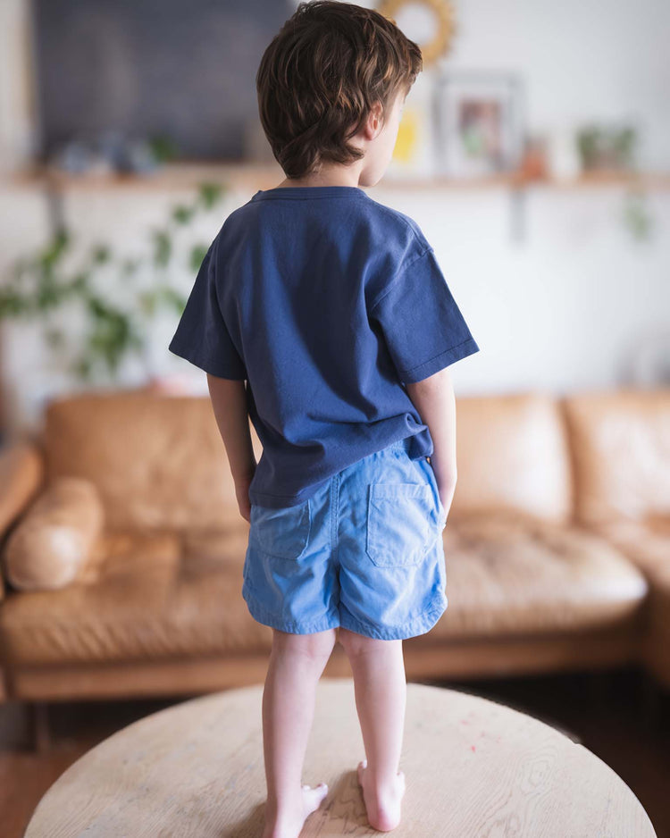 A young Sunchild with brown hair stands barefoot on a wooden table, wearing the Sunchild kazan tee in obscur and light blue shorts. The child faces away from the camera, with a tan couch and plants in the background.