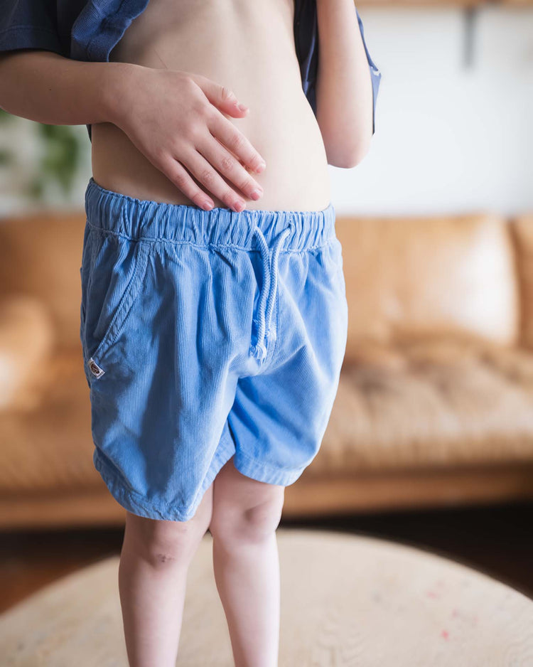 A child wears sunchild's hunter shorts in mare, featuring a light blue hue and drawstring waistband, standing indoors with one hand on their bare stomach. The blurred background reveals a tan sofa; the child’s head and upper body are partially out of frame.