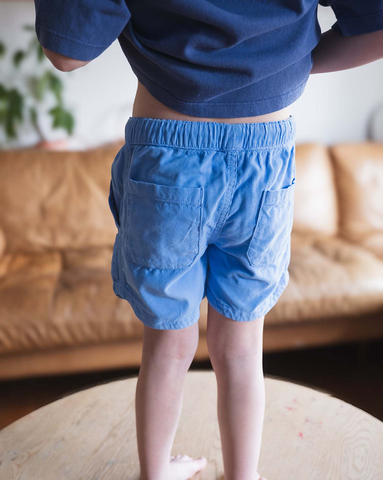 A child, seen from behind, stands barefoot on a wooden table wearing a navy shirt and Sunchild’s hunter shorts in mare with a drawstring waistband. A tan sofa and houseplant are blurred in the background.
